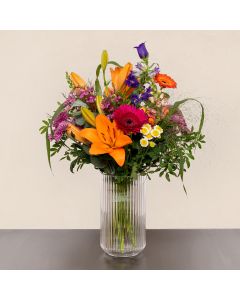 Buket med orange liljer, pink og lilla blomster i en klar glasvase på et bord.