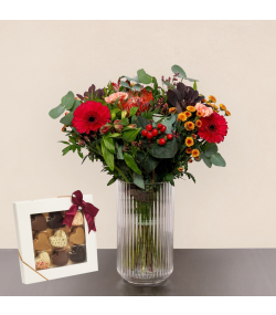 Buket med røde gerbera, orange blomster, røde bær og grønne blade i glasvase samt æske med chokolade.
