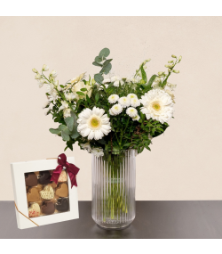 Nordisk blomsterbuket med hvide gerbera og hvide blomster i glasvase samt æske med chokolade.