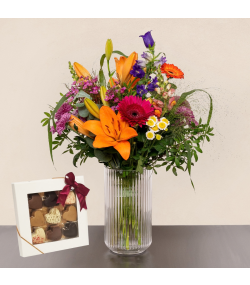 Buket med orange liljer og pink, lilla og hvide blomster i glasvase samt æske med chokolade.