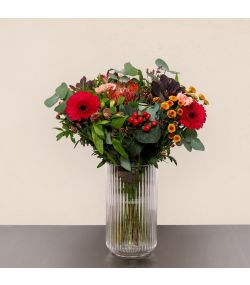 Buket med røde gerbera, røde bær, orange blomster og grønt i høj glasvase.