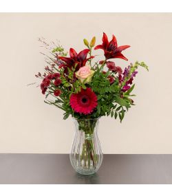 Buket med mørkerøde liljer, pink gerbera, lyserød rose og grønt i glasvase.