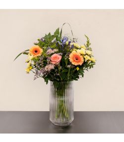 Pastel blomsterbuket med ferskenfarvede gerbera, lyserød rose, blå blomster og grønt i glasvase.