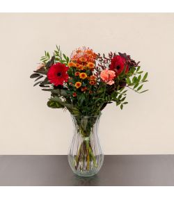 Buket med røde gerbera, orange blomster og lyserøde nelliker i glasvase.