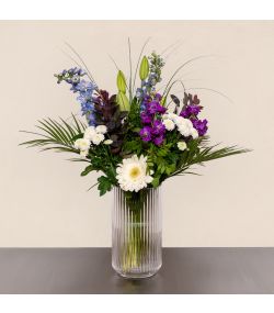 Trendy FLOWR buket med hvide gerbera, lilla blomster, liljeknopper og grønt i høj glasvase.
