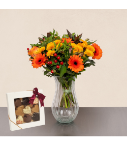 Efterårsbuket med orange gerbera, gule blomster, røde bær og chokolade i glasvase.