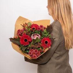 Ugens FLOWR buket med røde gerbera, rosa blomster, røde bær og grønt pakket i brunt papir.