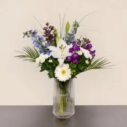 Trendy FLOWR buket med hvide gerbera, lilla blomster, liljeknopper og grønt i høj glasvase.